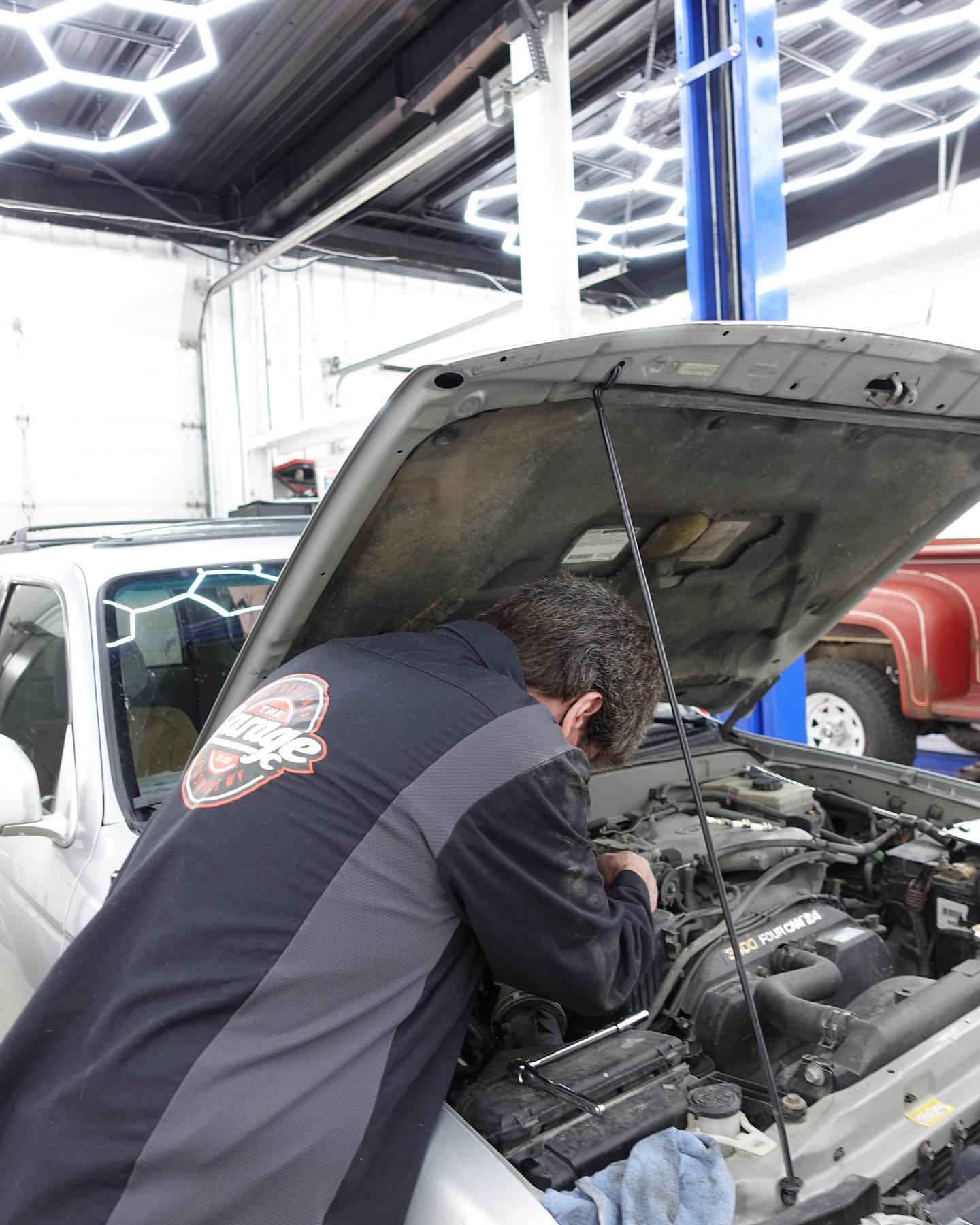 The Garage technician working under the hood of a light-colored SUV in the service bay